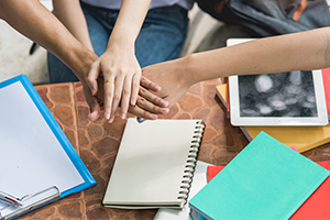 Hands stacked on top of each other above a table with notebooks, a tablet and school supplies on it